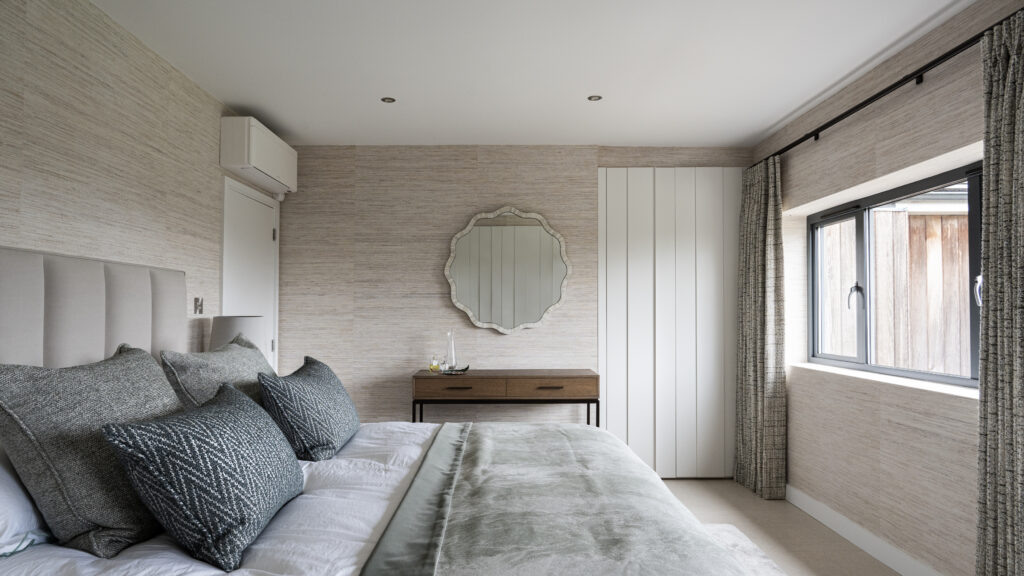 Bedroom with grasscloth wallpaper, round mirror above a console bench and a bed dressed in grey textured throws