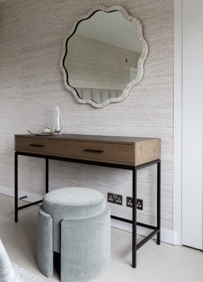 Dressing area with a scalloped round mirror, slim wooden desk and upholstered stool against textured wallpaper